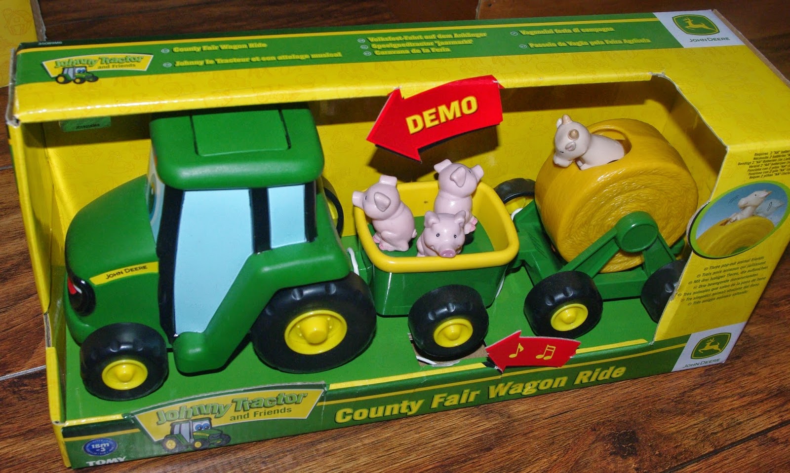 A Tomy John Deere toys set in a box featuring a green tractor with a yellow wagon carrying three pig figures and a trailer with a yellow hay bale. The packaging reads "Johnny Tractor and Friends County Fair Wagon Ride.