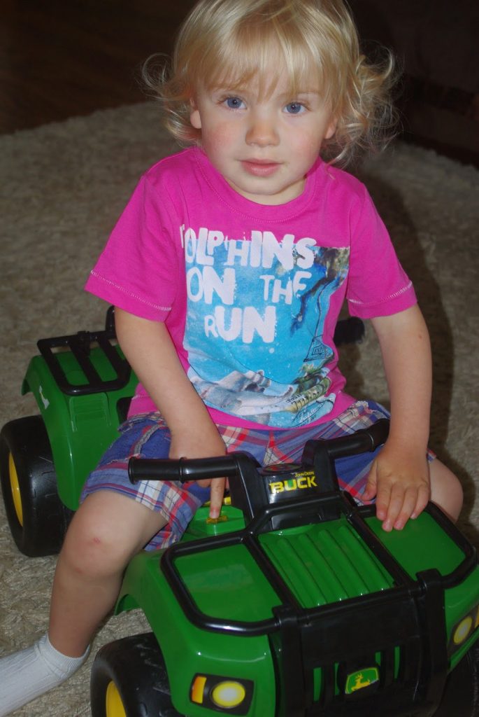 A young child with blonde hair sits on a green Tomy John Deere toy tractor indoors, wearing a bright pink "Dolphins on the Run" t-shirt and checked shorts, looking at the camera.