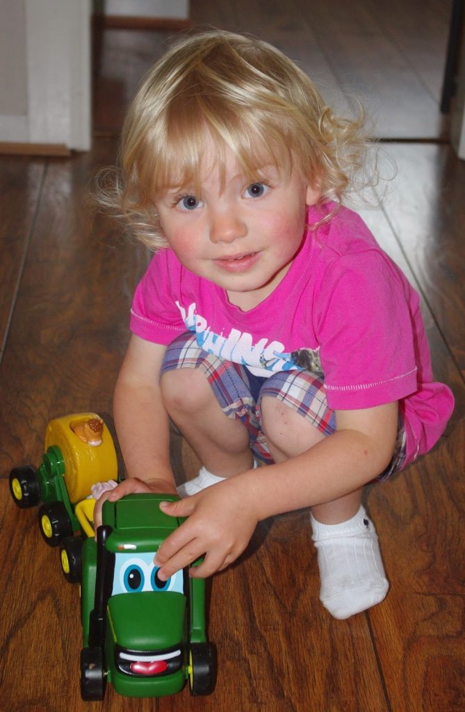 A young child with blond hair, wearing a pink shirt, checked shorts, and white socks, crouches on a wooden floor, smiling and playing with a green Tomy John Deere toy tractor that has a friendly face.