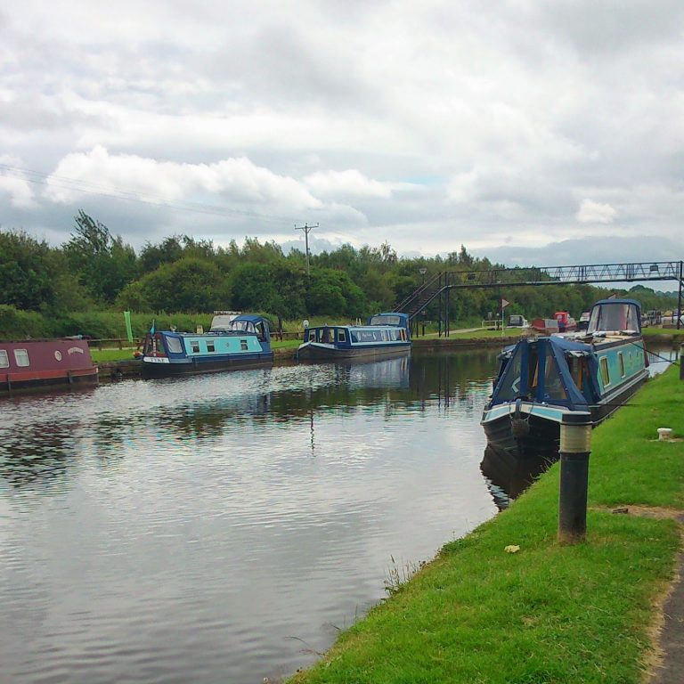 Colourful canal boats are moored along a calm canal, with green grass and trees on the banks under a cloudy sky. A footbridge crosses the canal in the background, creating the perfect scene for a Silent Sunday stroll.