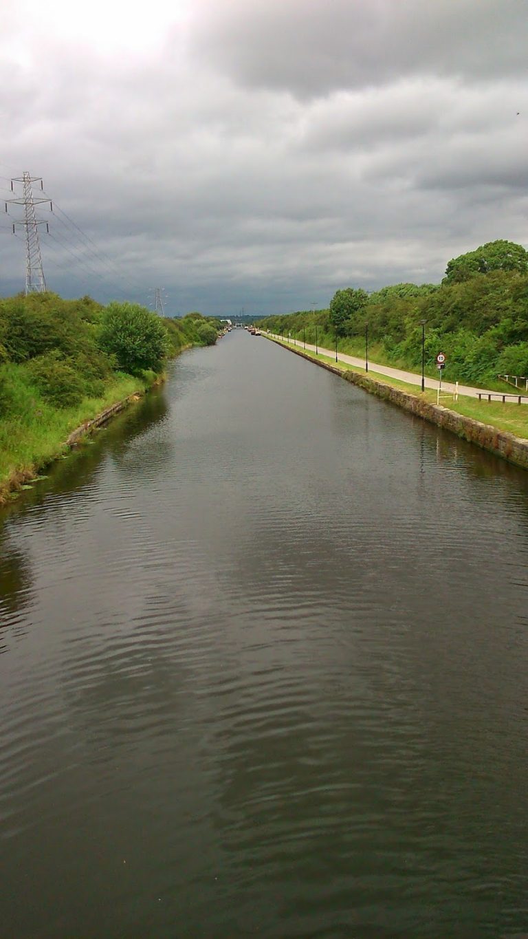 A calm canal stretches into the distance, bordered by green grass and trees on both sides under a cloudy sky. On this Silent Sunday, a paved path runs alongside the right bank with lamp-posts and a power line visible to the left.