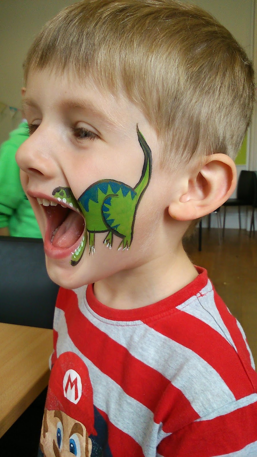 A young boy with short blond hair and a red-and-white striped shirt smiles with his mouth open on Silent Sunday, showing a green dinosaur face painting on his cheek, so the dinosaur’s mouth aligns perfectly with his.