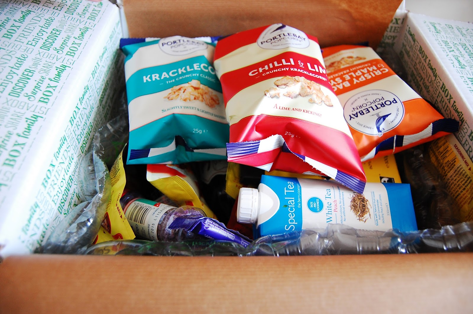 An open Degustabox snack subscription box filled with assorted packaged snacks, including packets of Kracklecorn, Chilli & Lime crisps, and a box of special tea, all surrounded by green and white packing material.