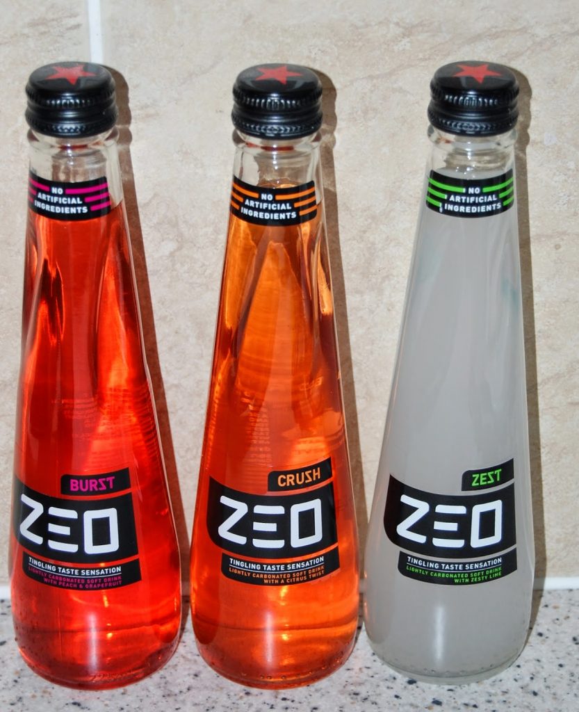 Three bottles of ZEO soft drinks—Burst, Crush, and Zest—are displayed side by side on a worktop. Each bottle boasts a black cap and "No Artificial Ingredients" label, perfect for discovering in your next Degustabox snack subscription box.