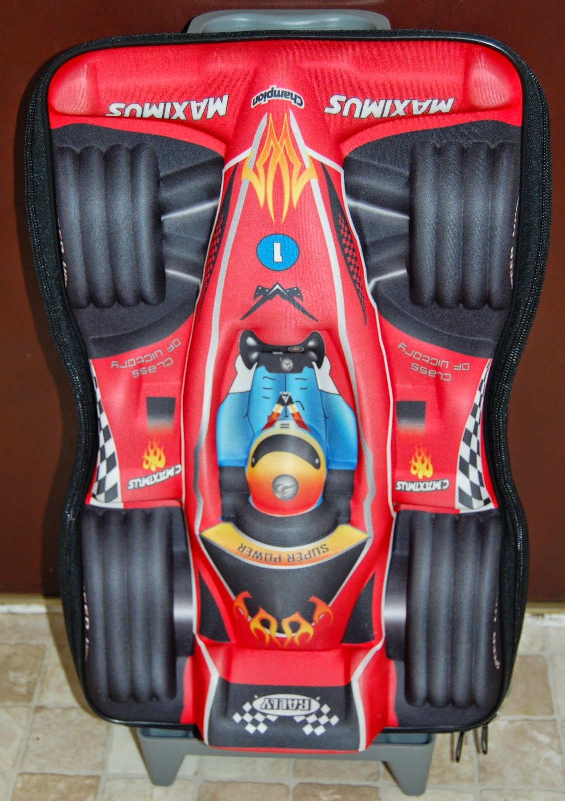 Travelling light with children is easy with this red suitcase designed to look like a racing car, featuring tyres, flames, race decals, and "MAXIMUS" on both sides. A cartoonish driver and detailed racing graphics complete the playful look.