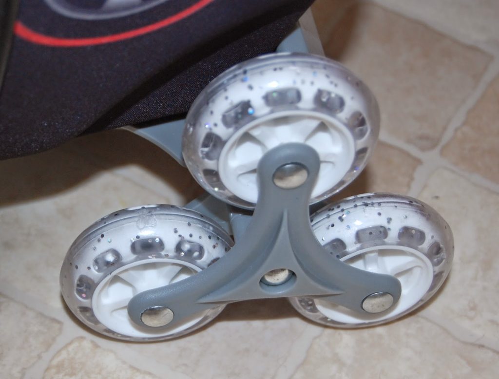 Close-up of three white wheels with grey spokes arranged in a triangular formation on a tiled floor, likely part of luggage designed for travelling light with children and easy rolling over steps.