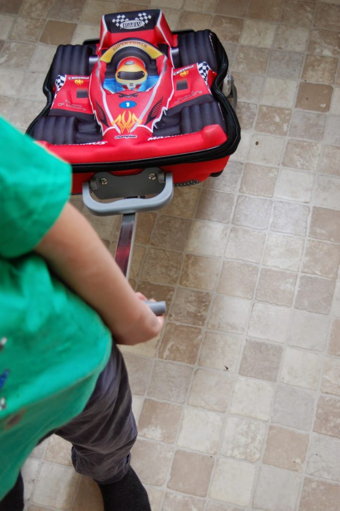Travelling light with children, a child in a green shirt and grey trousers pulls a red racing car suitcase by its handle across the tiled floor.
