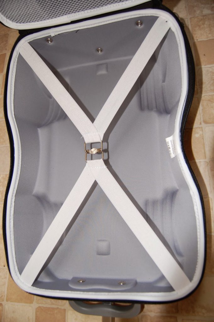 An empty, open suitcase lies on a tiled floor, showing a light grey interior with white elastic cross straps—perfect for travelling light with children—and a metal clasp in the centre to secure items.
