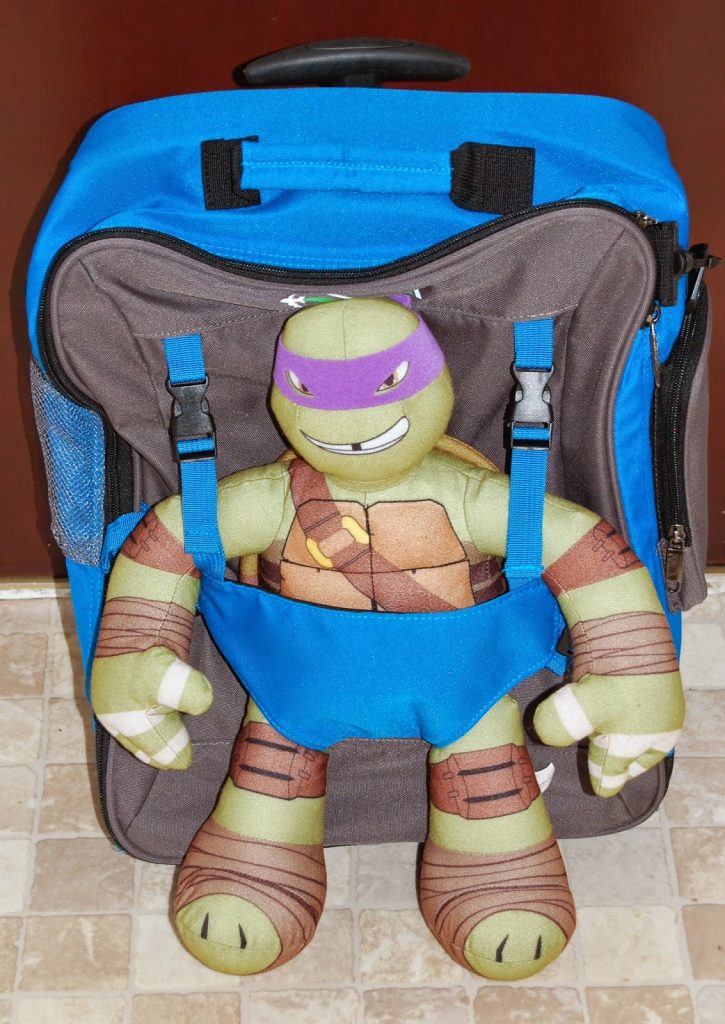 A plush Teenage Mutant Ninja Turtle toy with a purple mask is tucked into the front pocket of a grey and blue rucksack—a perfect example of travelling light with children—resting on a tiled floor in front of a brown door.