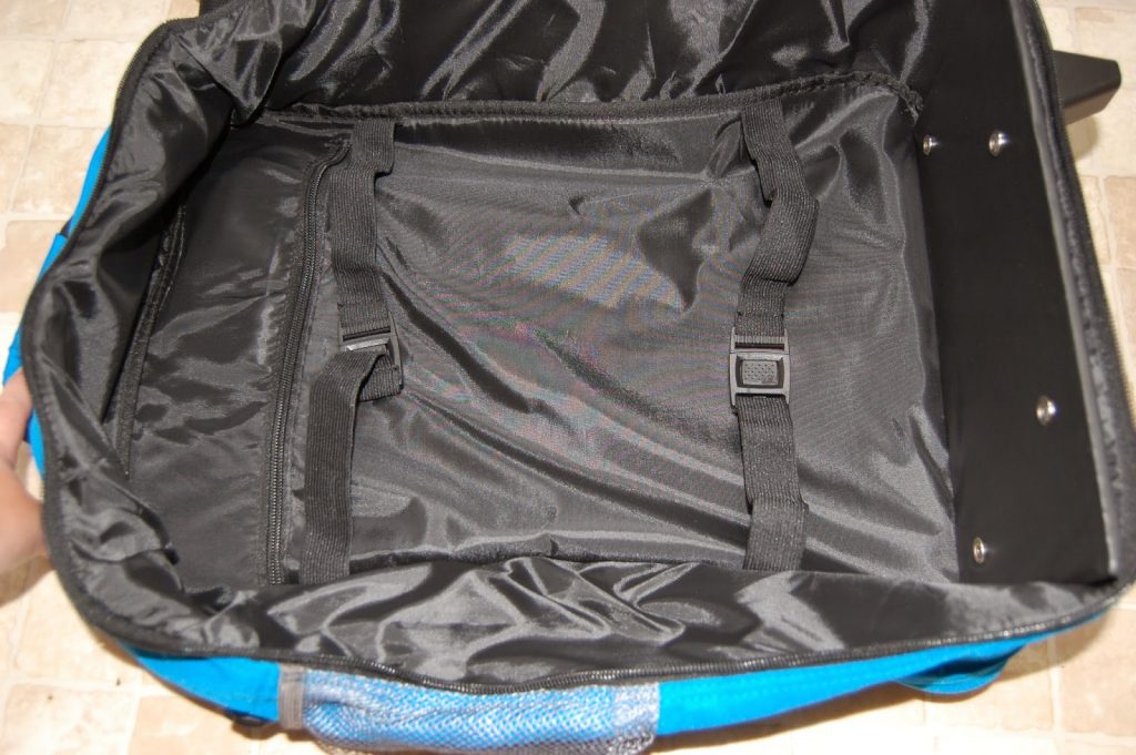 Open, empty black fabric interior of a luggage bag with adjustable straps and buckles, ideal for travelling light with children. Blue sides are partially visible at the edges, and the bag rests on a tiled floor.