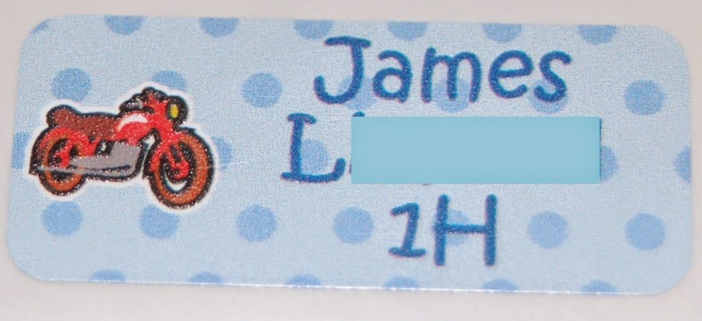 A rectangular name tag with blue polka dots, a red motorbike illustration on the left, and the text “James L—— 1H” on the right. Perfect for Back to School, part of the surname is obscured by a blue rectangle.