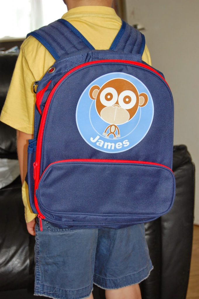 A child wearing a yellow shirt and blue shorts stands with their back to the camera, ready for the return to school, carrying a blue rucksack with a red zip and a round monkey patch labelled "James.