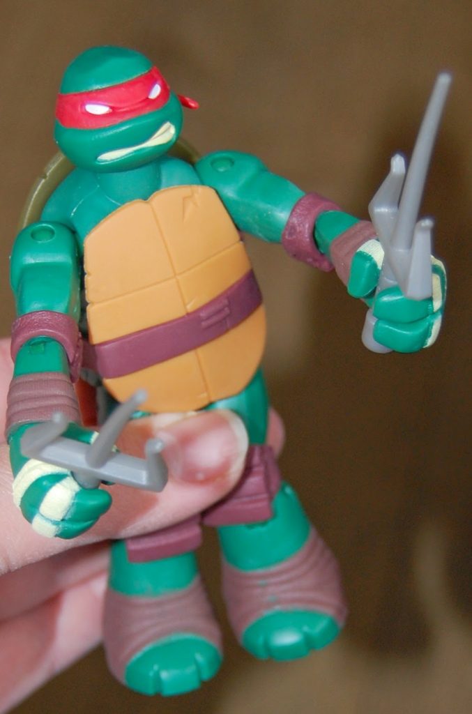 A hand holds an action figure of Raphael from Teenage Mutant Ninja Turtles Z-Line Ninjas, featuring a red mask, green body, and two grey sai weapons.