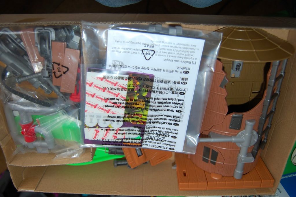 A cardboard box contains unassembled plastic toy parts, including a brown windmill structure and various grey and green components, resembling pieces from the Teenage Mutant Ninja Turtles Z-Line Ninjas playset, all packaged in clear plastic bags with instruction sheets on top.
