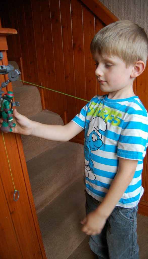 A young boy in a blue and white striped Smurfs T-shirt plays with a Teenage Mutant Ninja Turtles action figure attached to a string pulley system, inspired by the Z-Line Ninjas Zipline Playset, on a wooden staircase.