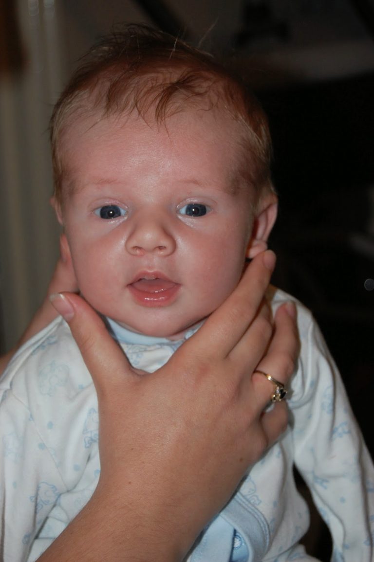 Flashback Friday: A baby with blue eyes and fair hair is gently held by an adult hand under the chin, wearing a light-coloured outfit and looking towards the camera.