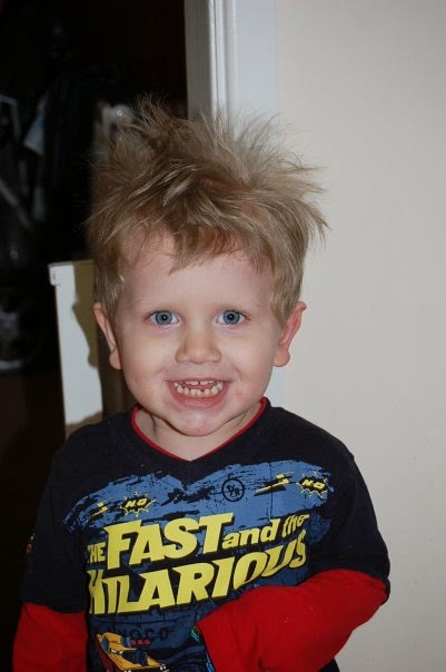 Smiling young child with messy blonde hair, wearing a navy and yellow "The Fast and the Hilarious" shirt, stands indoors against a cream-coloured wall for a perfect Flashback Friday moment.