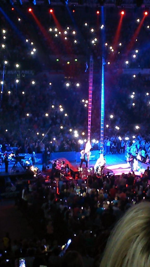 A brightly lit circus arena filled with a large audience; Ant and Dec stand near tall ladders at the centre, while the crowd surrounds them, many holding up glowing lights.