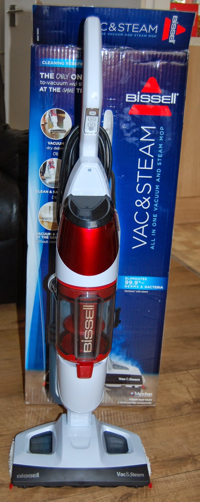 A Bissell Vac and Steam 2 in 1 Cleaner stands in front of its blue packaging box on a wooden floor. The white and red machine features the brand name clearly visible on the front.