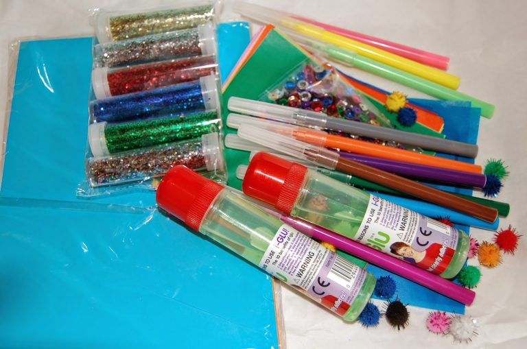 Assorted craft supplies including coloured tissue paper, glitter, glue, markers, pom-poms, sequins, and decorative gems—perfect for creating boy's dream homes—are arranged on a white surface.