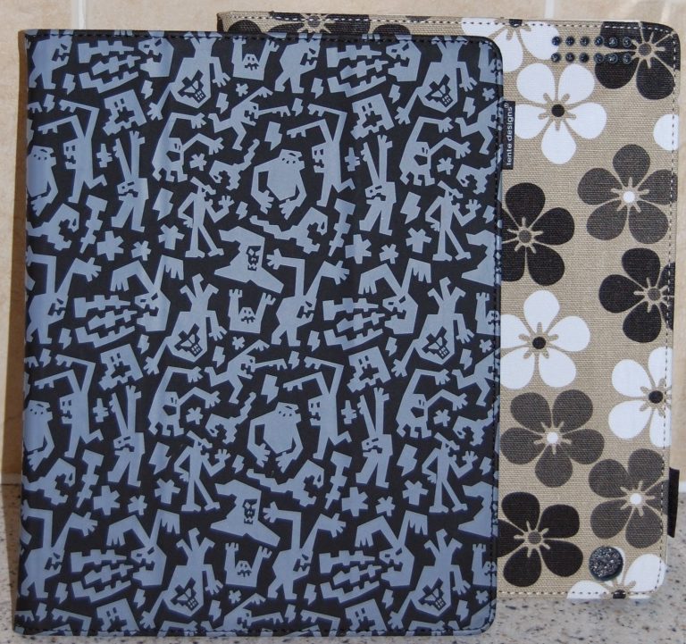 Two iPad accessories are standing upright. The front cover features a blue and black abstract pattern with geometric shapes, whilst the back cover displays a beige background adorned with large black and white flowers.