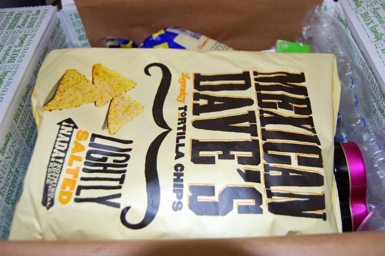 A bag of Mexican Dave's Lightly Salted Legendary Tortilla Chips inside a cardboard box, surrounded by packing materials and other grocery items from a snack subscription box.