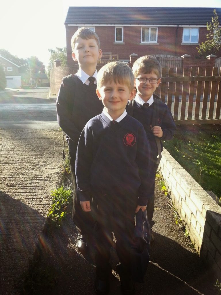 Three young boys in dark school uniforms stand smiling outside on a sunny Silent Sunday, with houses and a wooden fence in the background. Sunlight shines over them, creating a warm, cheerful atmosphere.