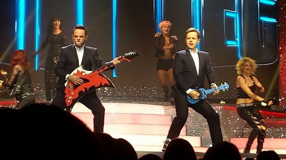 Ant and Dec, dressed in suits, pretend to play toy guitars on stage, surrounded by energetic dancers in black outfits, with bright lights and a glittery staircase in the background, creating a lively performance scene.