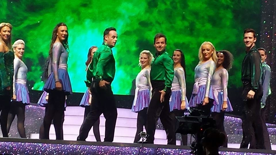 A group of dancers in shiny green and silver costumes perform on a stage with glowing green lights and steps in the background, reminiscent of an Ant and Dec spectacular.
