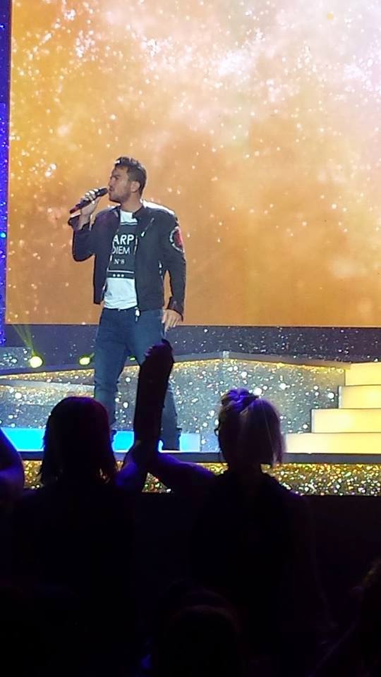 A singer performs on a brightly lit stage, holding a microphone. The background glows with warm golden lights, whilst silhouetted audience members—including Ant and Dec—watch from the foreground.