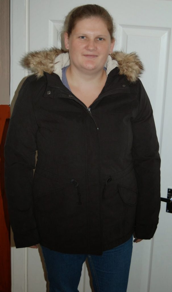 A woman stands indoors in front of a white door, wearing a black winter coat with a fur-trimmed hood and blue jeans. She is looking at the camera with a neutral expression, ready for the winter season.