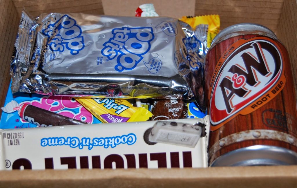 A box filled with assorted snacks including Pop-Tarts, a can of A&W root beer, a Hershey’s Cookies 'n' Creme bar, Tootsie Rolls, and colourful sweet packages—perfect for any Taffy Mail Review or British sweet subscription unboxing.