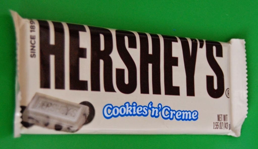 A Hershey's Cookies 'n' Creme chocolate bar in its white and blue wrapper, lying on a green surface—just the kind of treat you might find in a British sweet subscription like Taffy Mail. The wrapper shows the Hershey's logo and biscuit pieces.