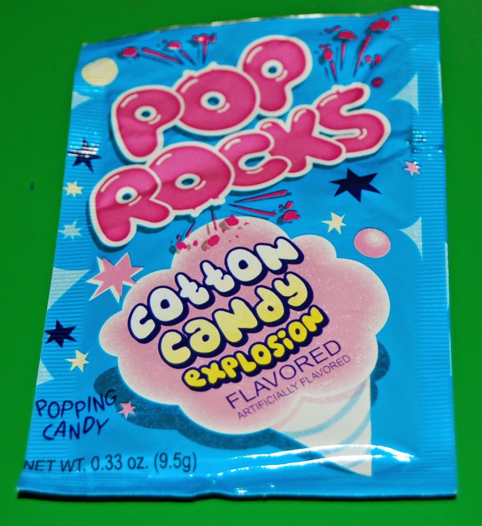 A blue packet of Pop Rocks popping candy, candyfloss explosion flavour, sits on a green background. Featured in our British sweet subscription, Taffy Mail review, the pack shows colourful stars and a pink candyfloss illustration.