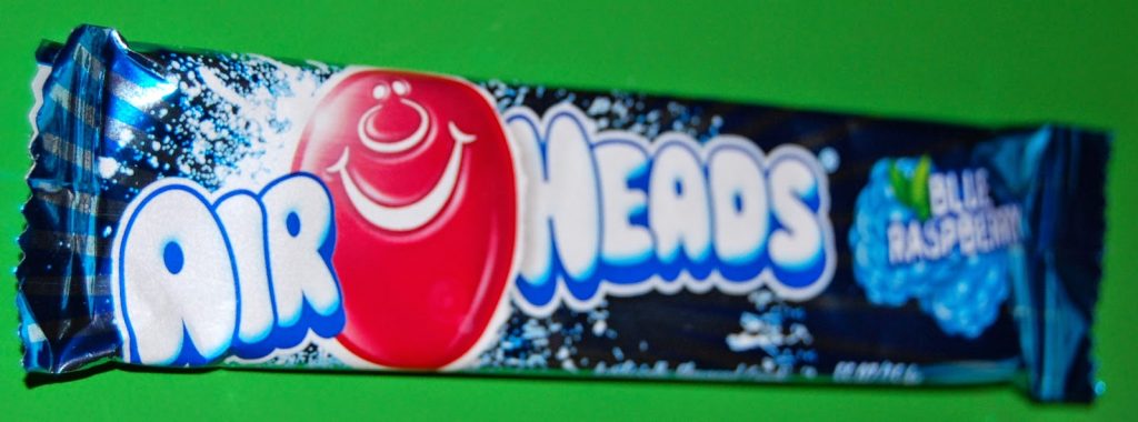 A blue raspberry AirHeads sweet in its shiny wrapper, as featured in Taffy Mail Review. This British sweet subscription highlights confectionery with red smiling face logos and bold, white “AirHeads” text on a green background.