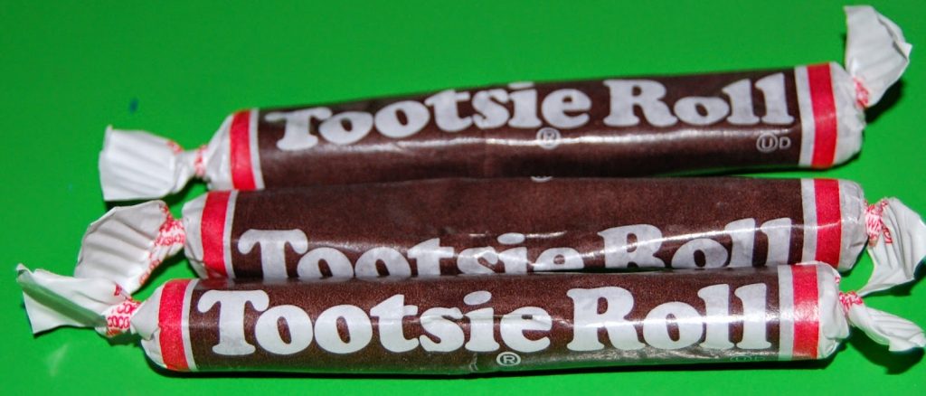 Three Tootsie Roll sweets wrapped in brown and white packaging are arranged side by side on a bright green background, perfect for a British sweet subscription like Taffy Mail.