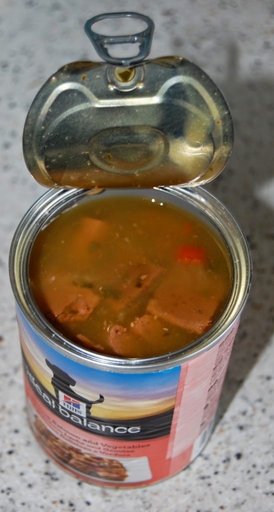 An open tin of Hills Ideal Balance Dog Food sits on a speckled worktop. The lid is partially pulled back, revealing chunks of meat in gravy. The label displays the brand name and additional text.
