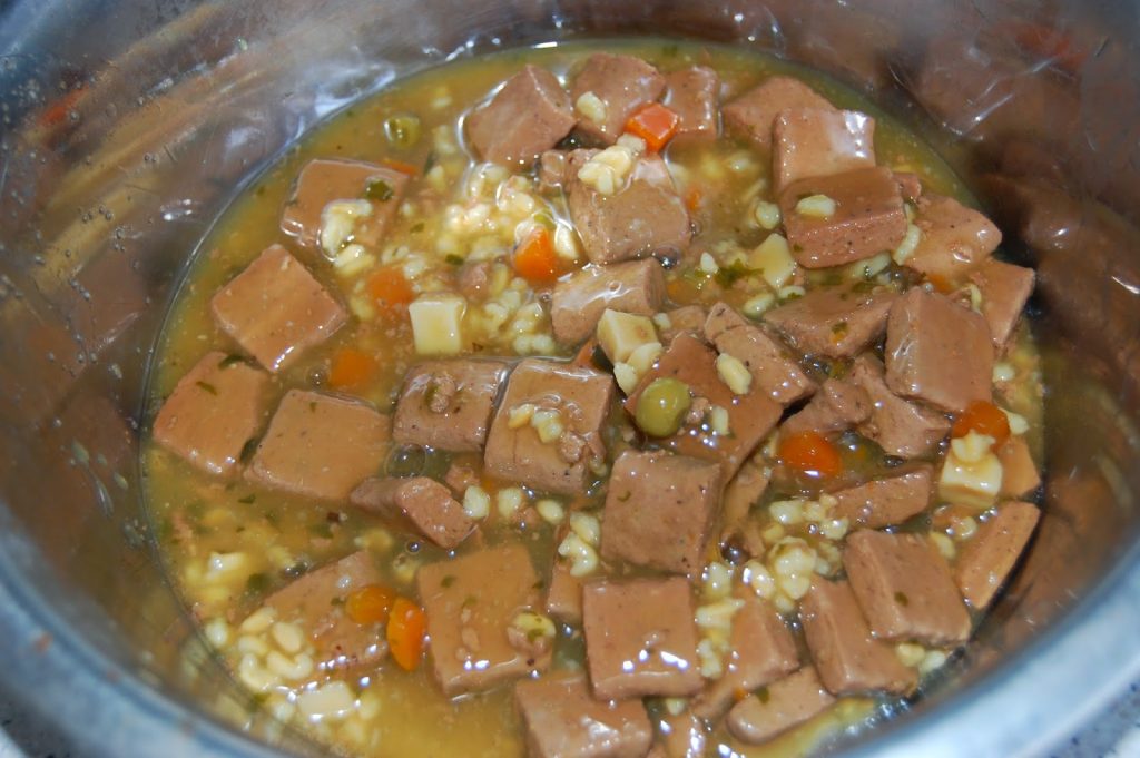Chunks of brown meat, peas, carrots, and grains in a thick, yellow-brown gravy inside a metal bowl, resembling a serving of Hills Ideal Balance Dog Food stew.