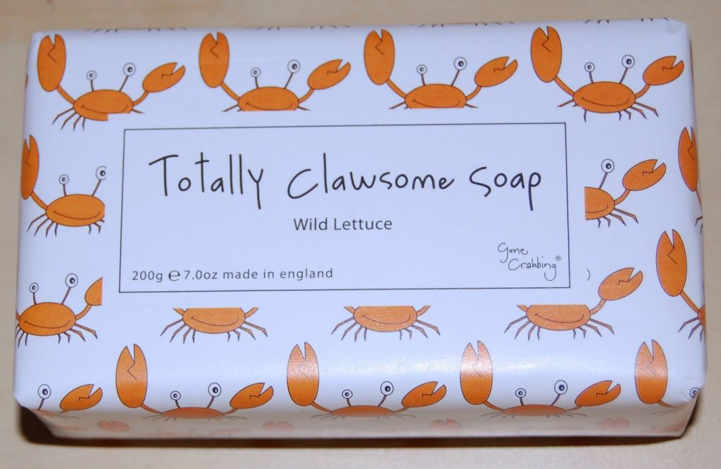 A white soap bar wrapped in paper decorated with orange cartoon crabs, including Colin the Crab. The label reads: "Totally Clawsome Soap, Wild Lettuce, 200g e 7.0oz made in England.