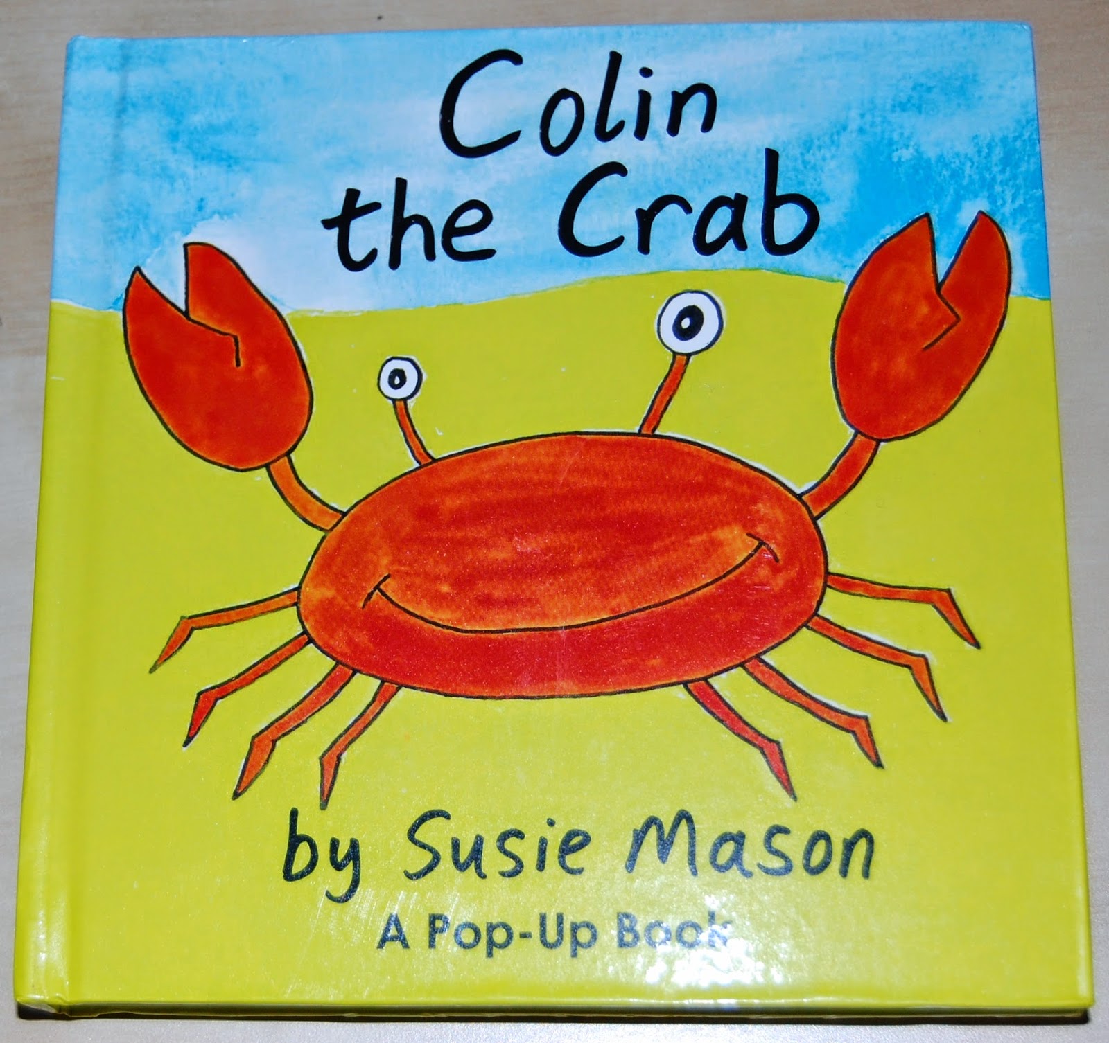 The cover of the children’s book "Colin the Crab" by Susie Mason features Colin the crab smiling with raised claws on a yellow and blue background, with the text "A Pop-Up Book" at the bottom.