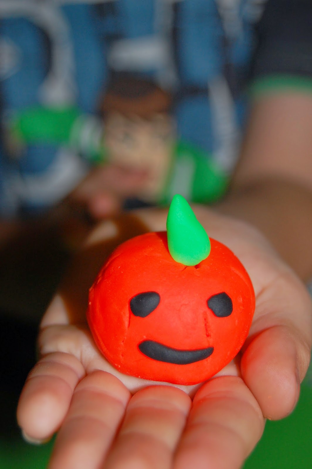 On this Silent Sunday, a child holds a small orange clay ball with a smiling face and green stalk, resembling a pumpkin, gently in their open hands. The background is softly blurred.