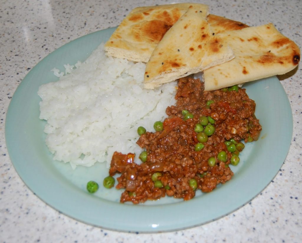 A plate with white rice, savoury Keema cooked with peas in a red sauce, and several pieces of naan bread on a speckled worktop—a delicious sample of Cook With Keema recipes.