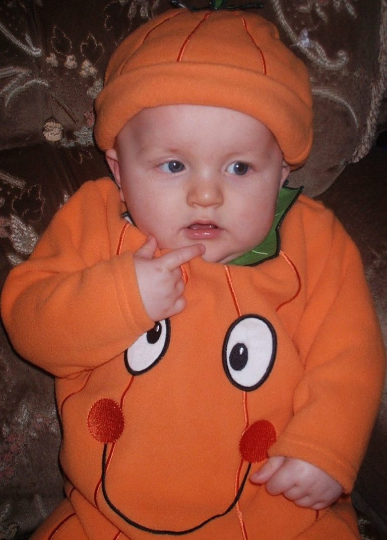 Flashback Friday: A baby wearing an orange pumpkin costume with a matching hat sits on a patterned brown chair, touching their face with one finger and looking slightly to the side.