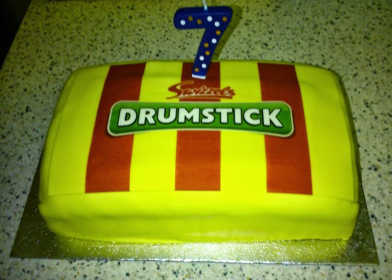 A rectangular cake decorated like a Drumstick sweet wrapper with red and yellow stripes, a "Drumstick" label, and a blue polka dot number 7 candle on top—perfect for a Silent Sunday celebration. The cake sits on a speckled worktop.
