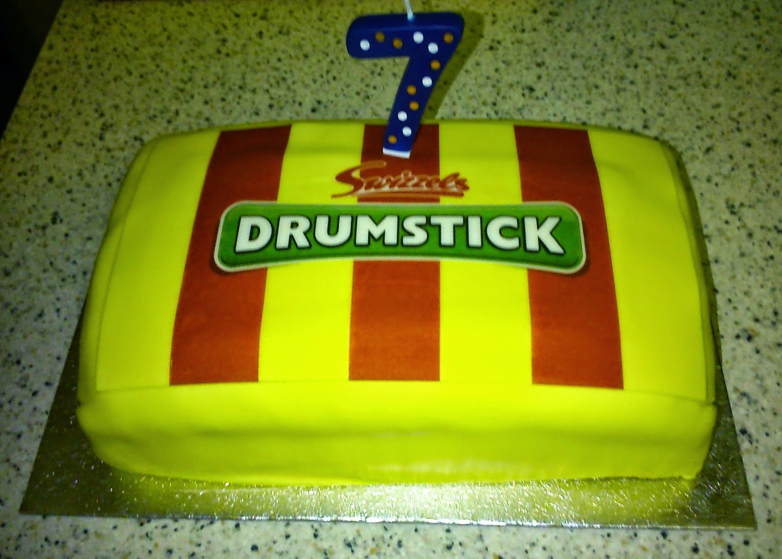 A rectangular cake decorated like a Drumstick sweet wrapper with red and yellow stripes, a "Drumstick" label, and a blue polka dot number 7 candle on top—perfect for a Silent Sunday celebration. The cake sits on a speckled worktop.