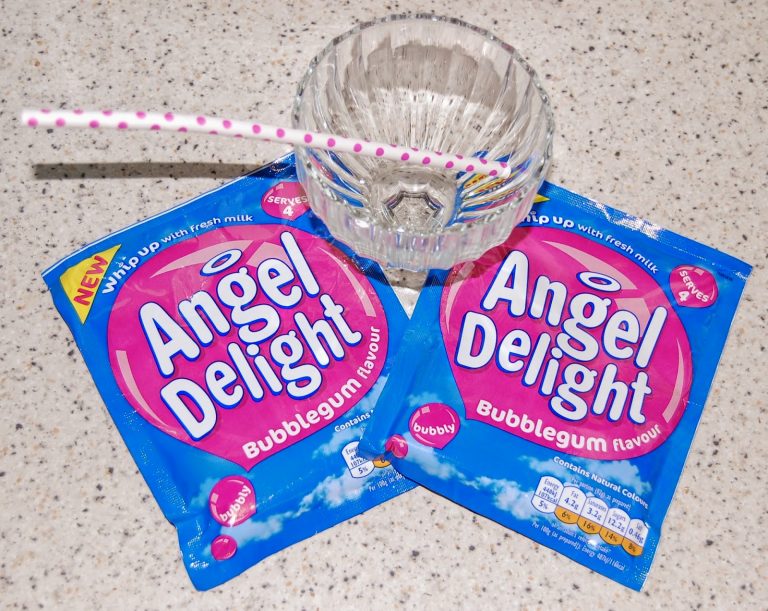 Two packets of Angel Delight Moments bubblegum flavour dessert are on a worktop next to an empty glass bowl with a pink spotty straw. The blue and pink packaging features a playful cloud design.