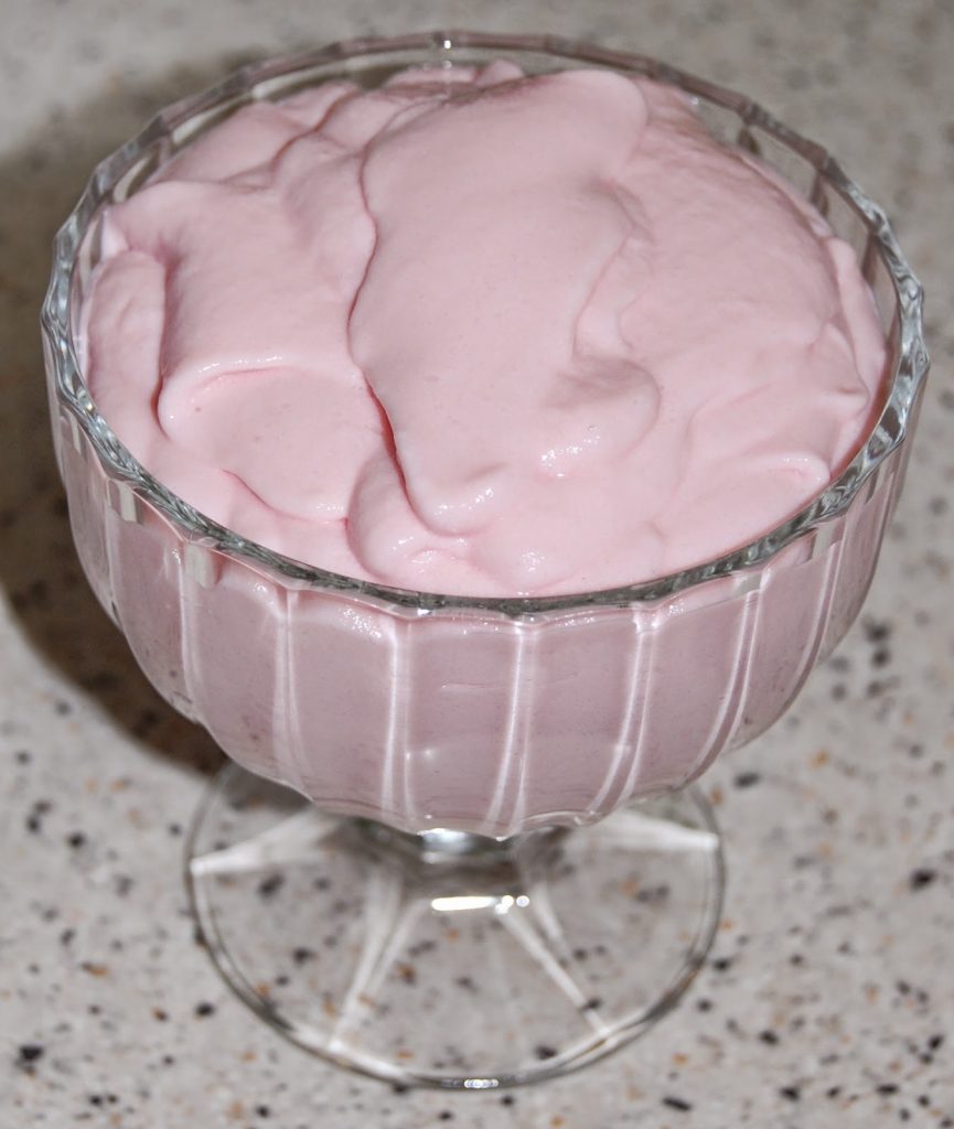 A glass dessert bowl filled with thick, creamy pink Angel Delight mousse sits on a speckled worktop.