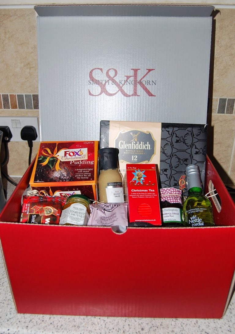 A red gift box filled with assorted food and drink items, perfect for a relaxing Silent Sunday, including mini bottles of alcohol, tea, pudding, jam, mustard, and sweets. The S&K logo is on the inside lid as the box sits on a kitchen worktop.