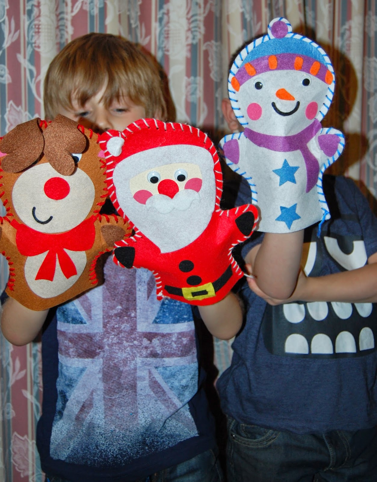 Two children hold up festive felt puppets— a reindeer, Father Christmas, and a snowman—during Silent Sunday. Their faces are partially hidden by the puppets, with a patterned curtain as the backdrop.