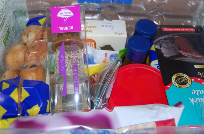 A selection of food items and meal kits, including muffins, bottled drinks, snacks, cutlery, and packaged meals, all neatly arranged inside a clear Degustabox subscription box.